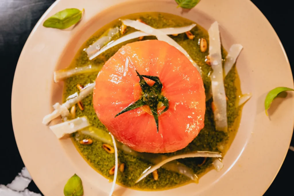 Tomate pelée farcie à la mozzarella di bufala sur un lit de pesto, avec copeaux de parmesan, pignons de pin et basilic