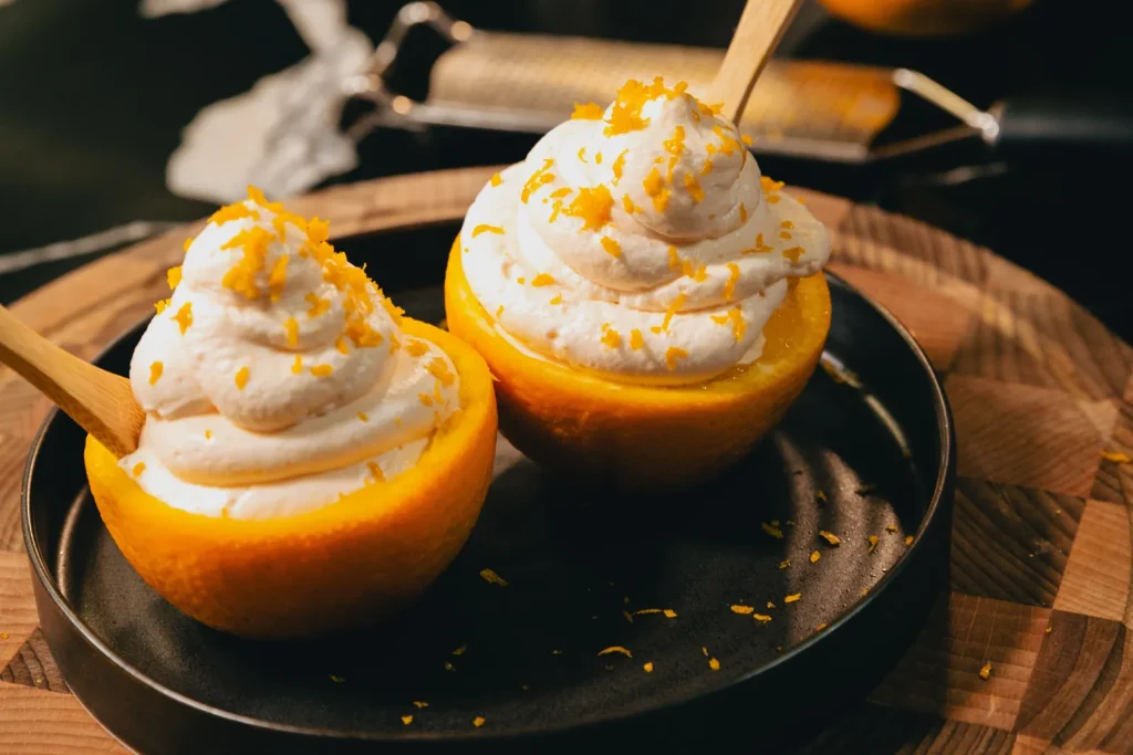 Deux demi-oranges garnies d'une mousse blanche au mascarpone et parsemées de zestes d'orange, sur une assiette noire